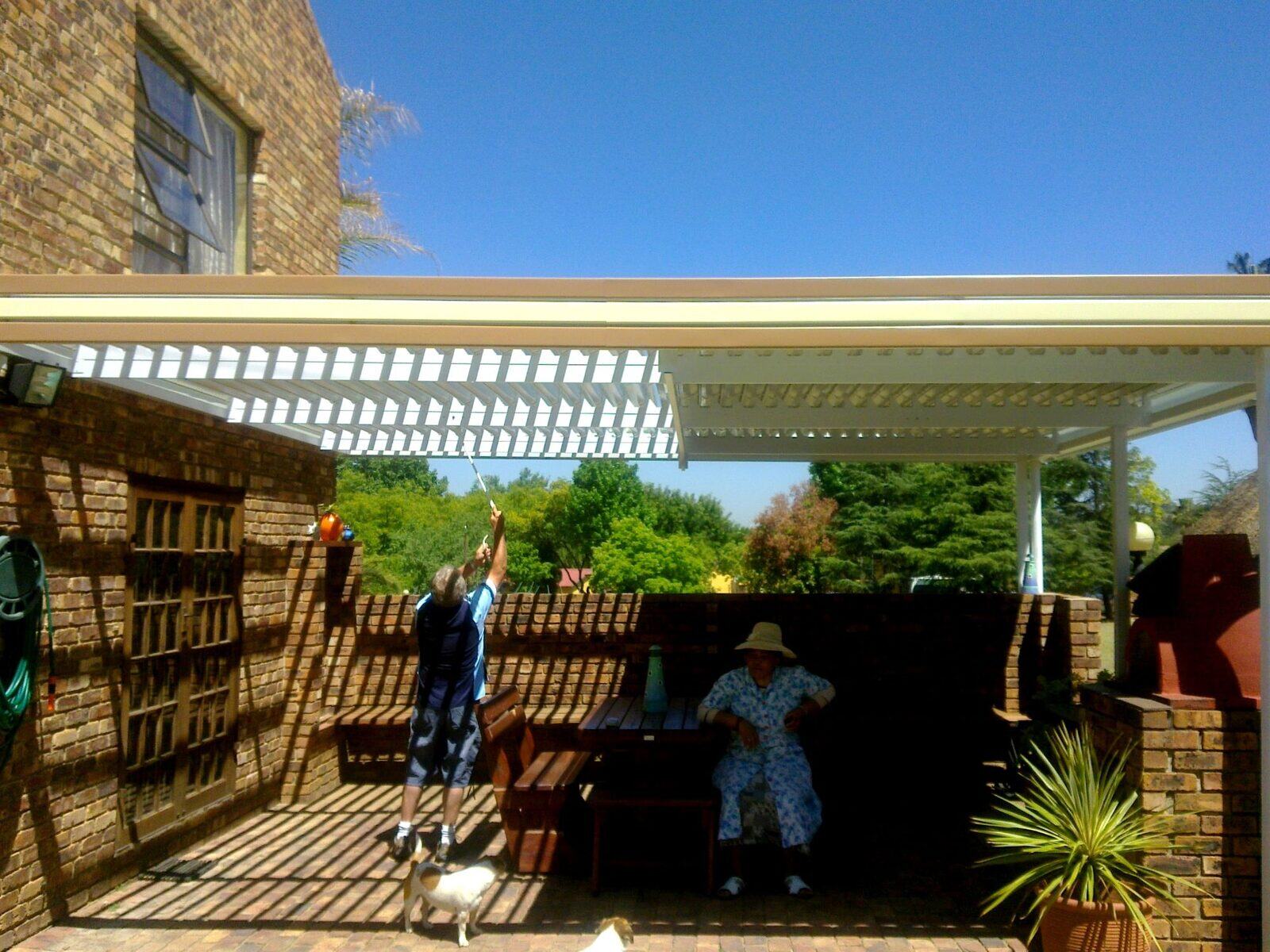 White patio cover with an adjustable louvre section being opened with a winding rod over a paved braai area