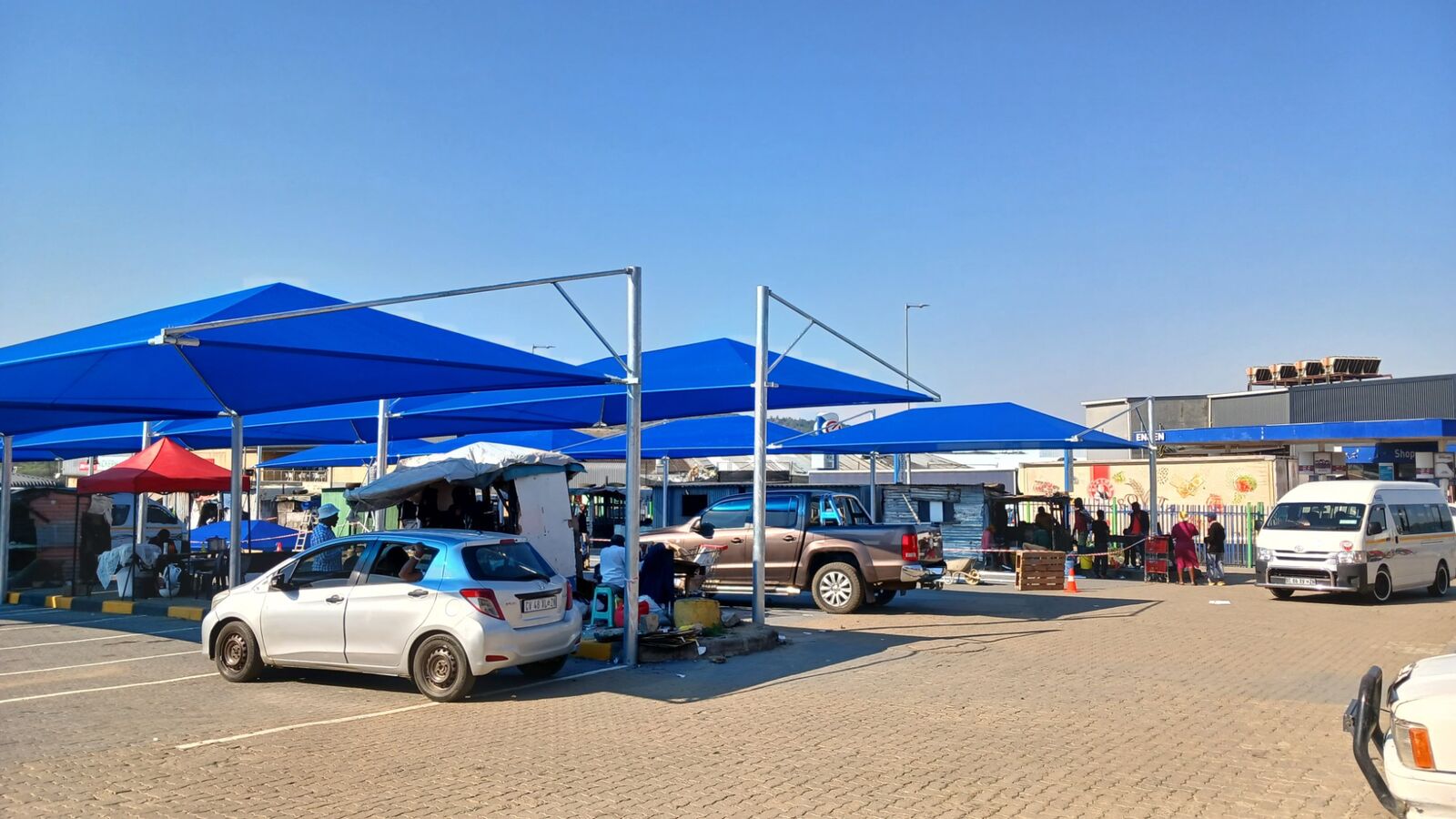 Row of blue shade-cloth shadeports on galvanised steel frames covering parking bays at a retail centre
