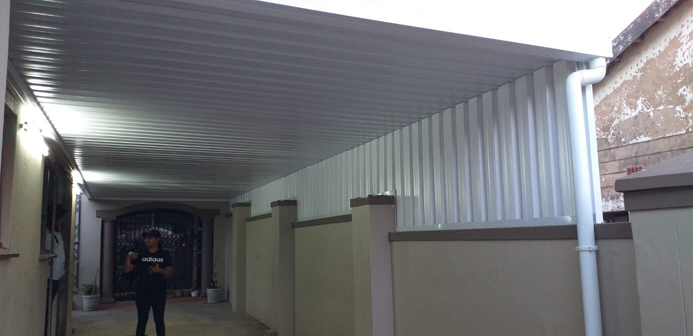 White IBR-roofed carport running the length of a narrow side driveway, with a boundary-wall gutter and downpipe